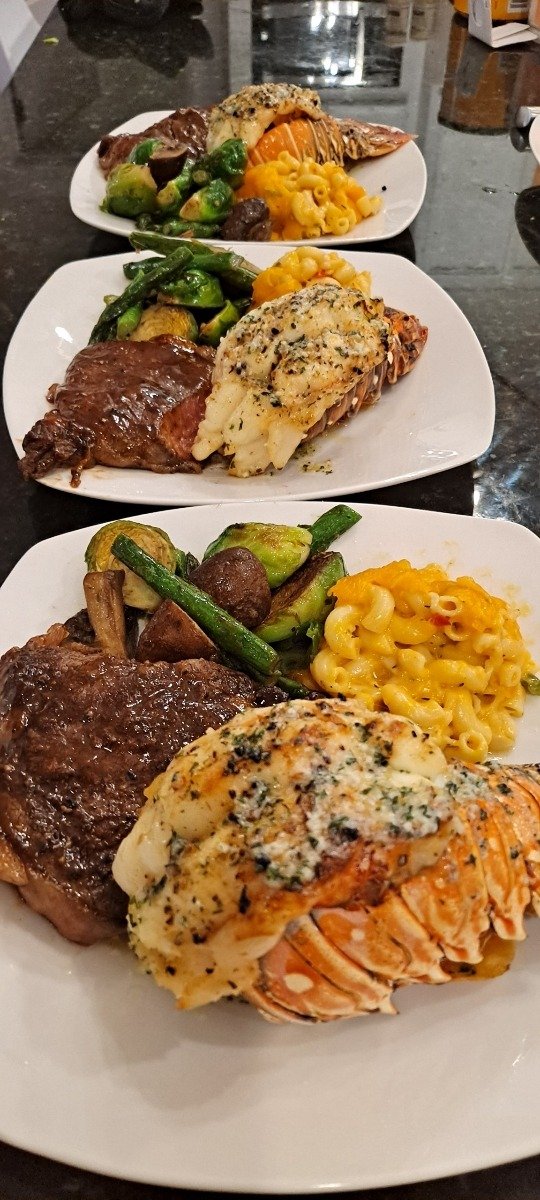Three plates of lobster tail, steak, macaroni and cheese and green vegetables lined up for service