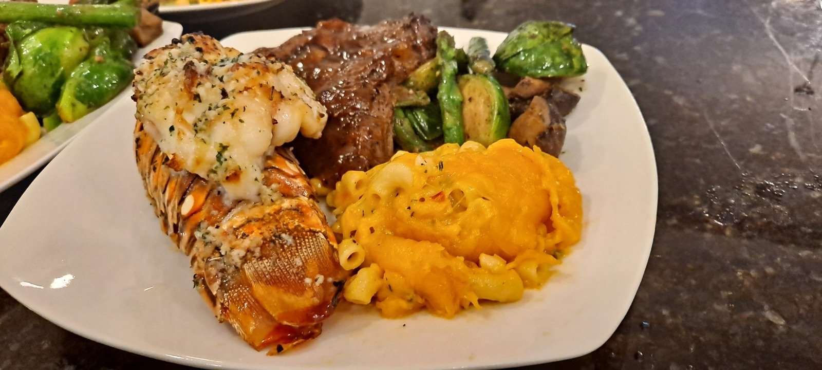 A plated lobster tail and sliced steak with macaroni and cheese and Brussels sprouts